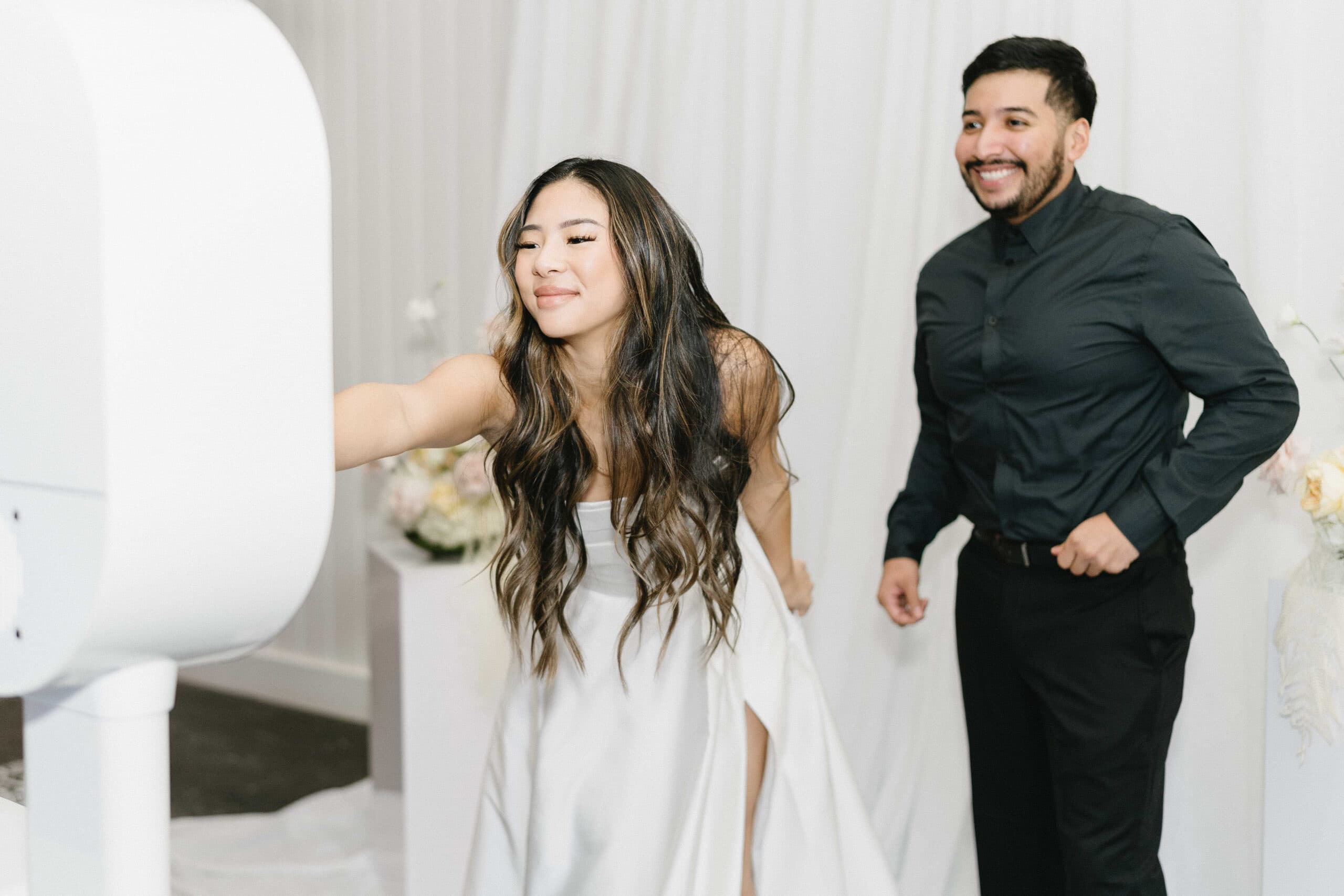 A bride reaches for the white booth screen while her guest waits for the frame