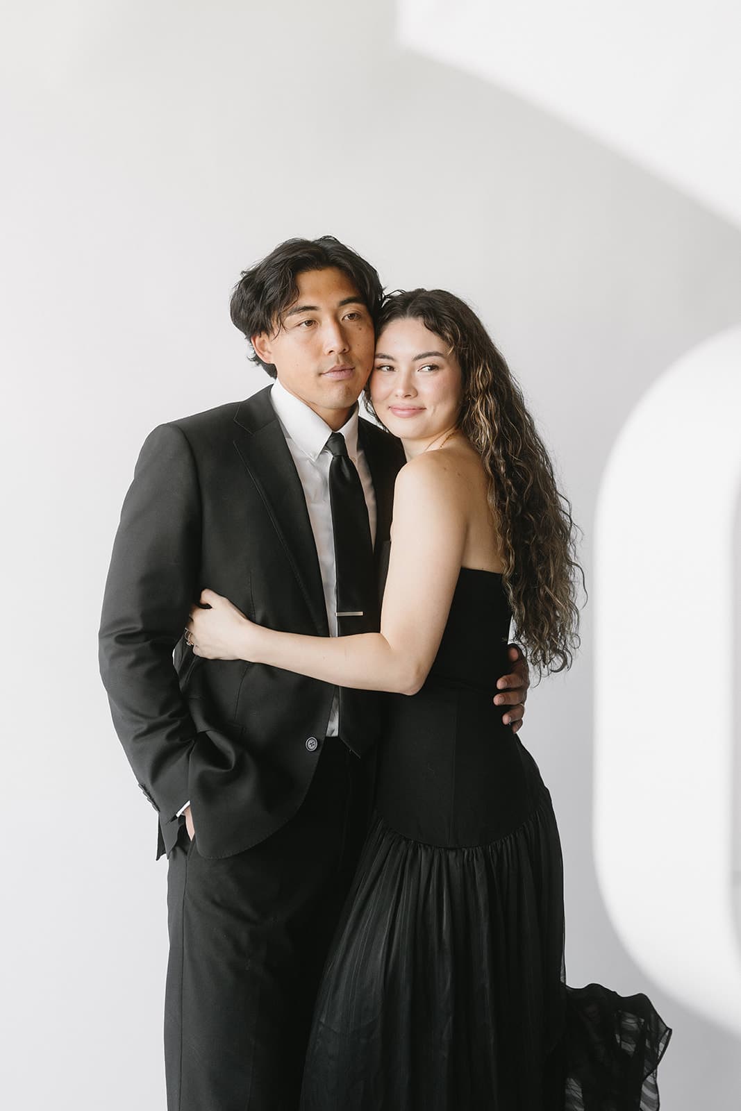 A couple in black-tie evening dress, editorial portrait against a bright studio wall